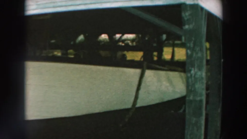 The boat remains in shadow under the wooden pier, with a vertical post and partial lawn visible beyond.