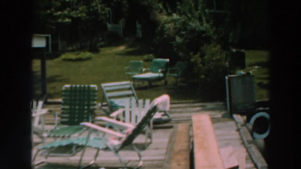 1964 Lake Winnebago frame showing mid-century lawn chairs on a deck with a grassy yard and shrubs beyond.