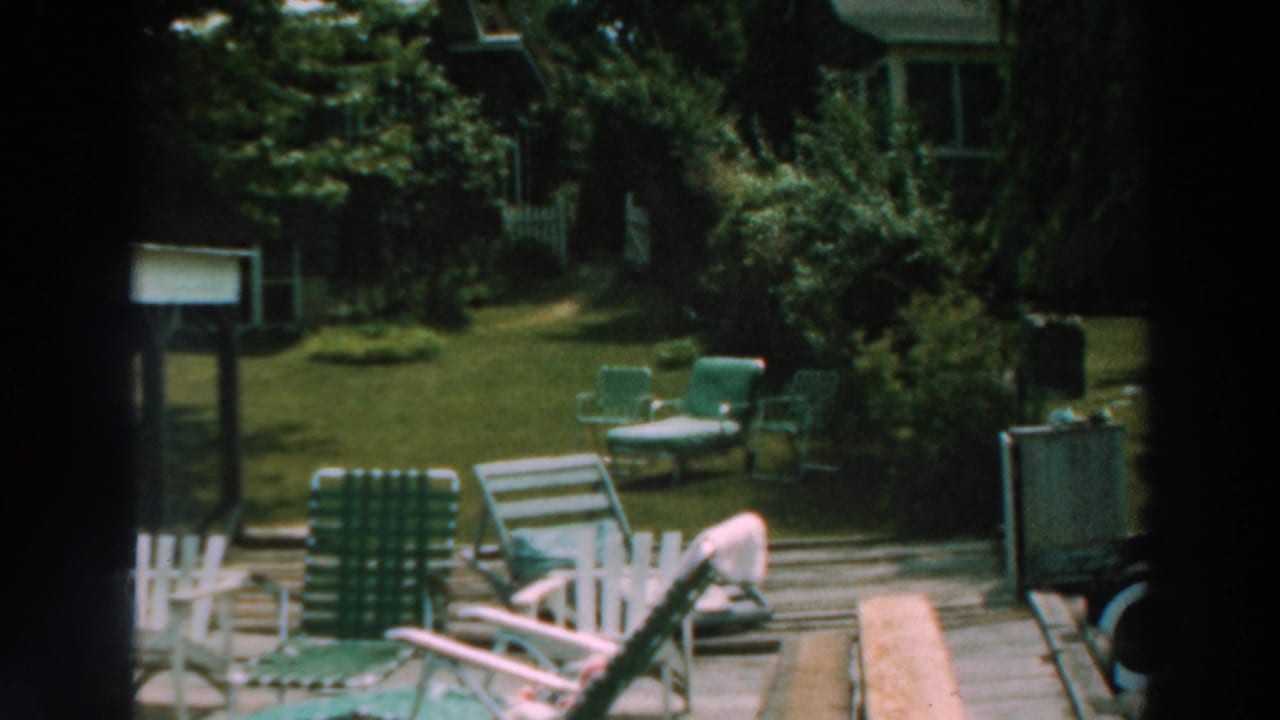 Green and white lawn chairs on a wooden deck beside a leafy yard.