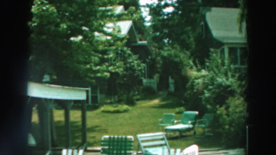 1964 Lake Winnebago yard view with green chairs, lawn, shrubs, trees, and a house exterior seen through dark framing.