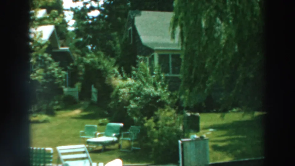 1964 Lake Winnebago home-movie view of a green lawn with patio chairs, trees, shrubs, and a house in soft focus.