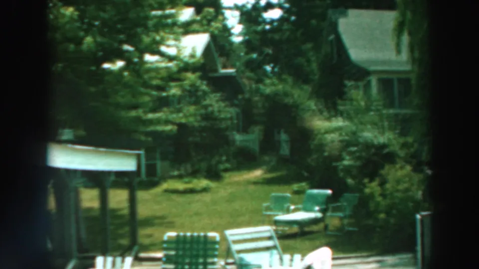 1964 Lake Winnebago frame showing a quiet green backyard with lawn chairs and a nearby house partly obscured by trees.