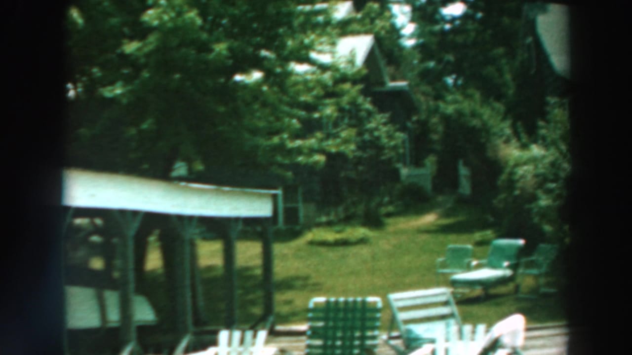 1964 Lake Winnebago home-movie frame showing a wooden dock, covered boat shelter, white boat, green lawn chairs, and trees.