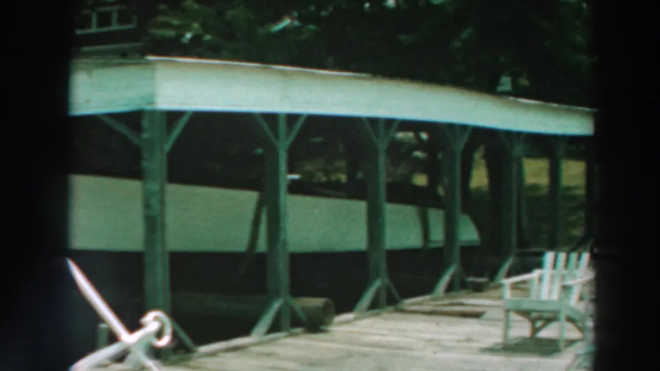 Closer 1964 lakeside view of the covered dock, white boat, support posts, wooden planks, and a chair near the shelter.