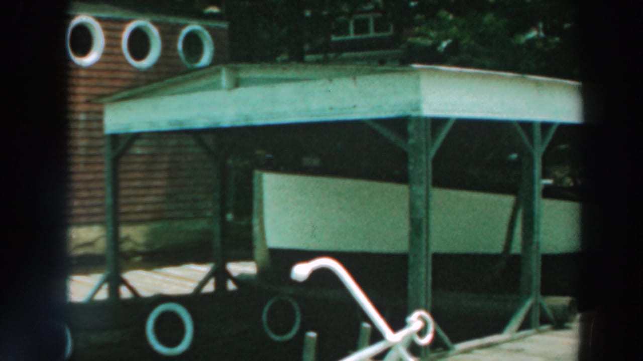 1964 Lake Winnebago home-movie frame showing a dark-sided house porch, white trim, circular windows, lawn, and garden details.