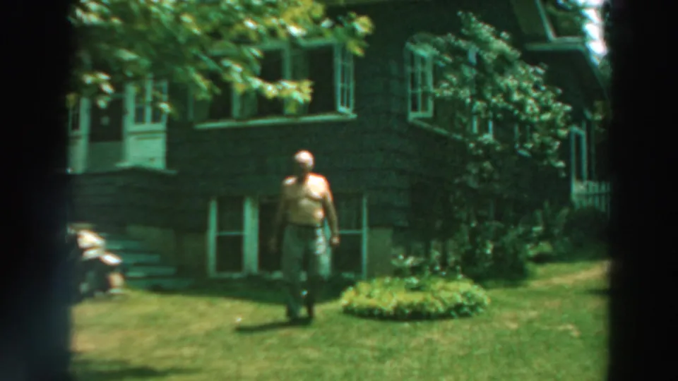 1964 Lake Winnebago frame of a shirtless man walking across a green lawn before a dark house with white-trimmed windows.