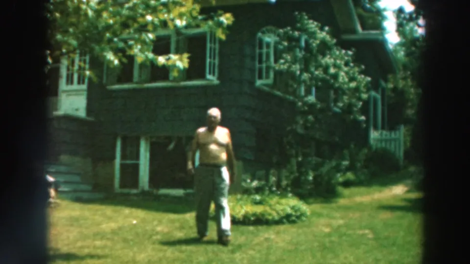 1964 Lake Winnebago home-movie frame with a shirtless man on the lawn, shrubs and trees around a dark residential house.
