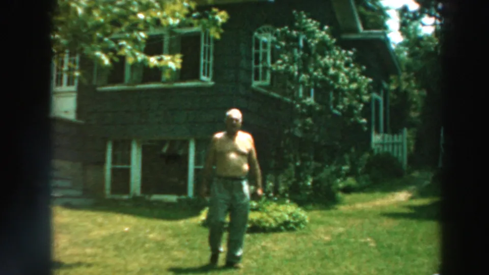 1964 Lake Winnebago frame showing a shirtless man approaching on the lawn, with a dark house, white windows, trees, and shrubs behind him.