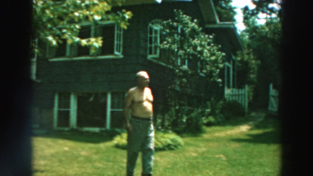 1964 Lake Winnebago home-movie frame showing a shirtless adult man on a sunlit lawn before a dark house and garden.