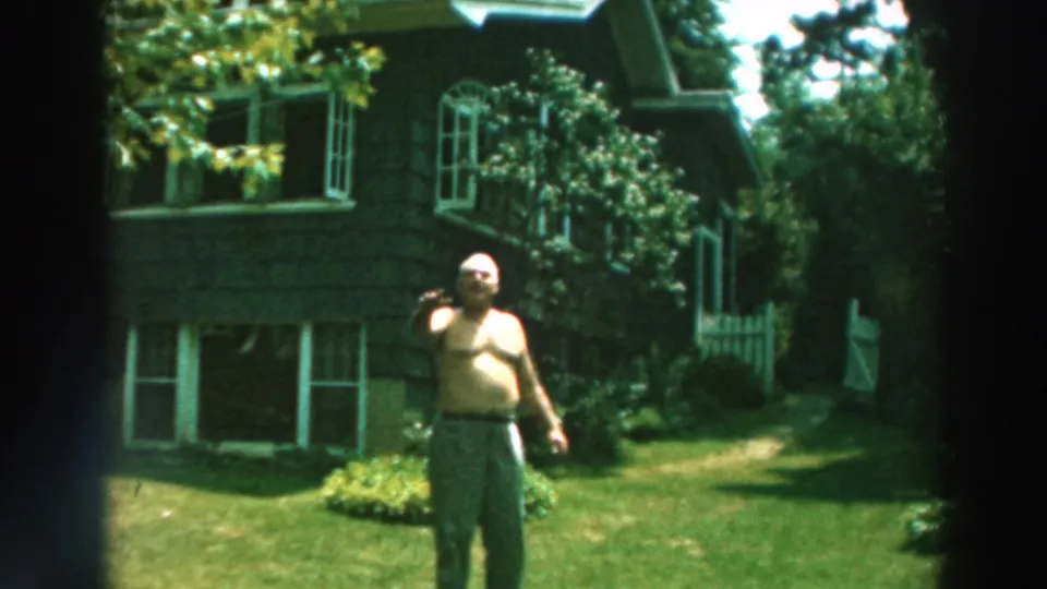 1964 Lake Winnebago frame with a shirtless man beginning to wave while standing on grass near a house and shrubs.