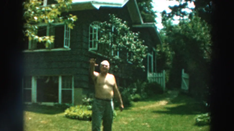1964 Lake Winnebago home-movie frame showing the man waving toward the camera in front of a house and greenery.
