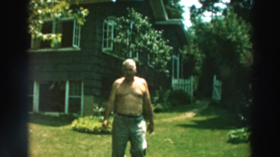 1964 Lake Winnebago frame showing the man walking past the camera-side view across a green yard by a dark-shingled house.