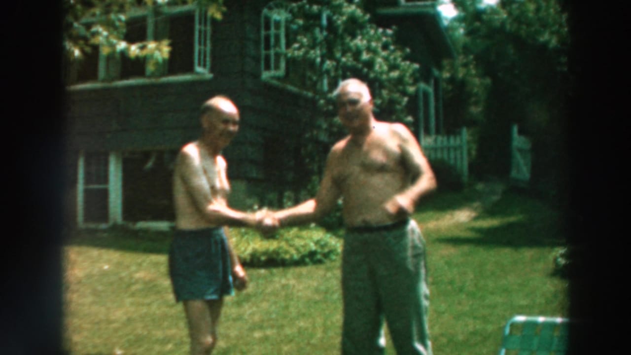 Two older shirtless men shake hands on a lawn in front of a house.