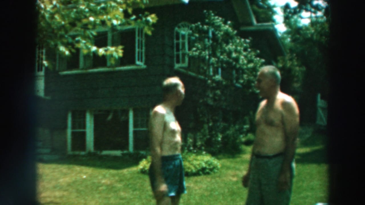 1964 Lake Winnebago home-movie frame showing two shirtless men greeting on a lawn beside a dark-sided house.