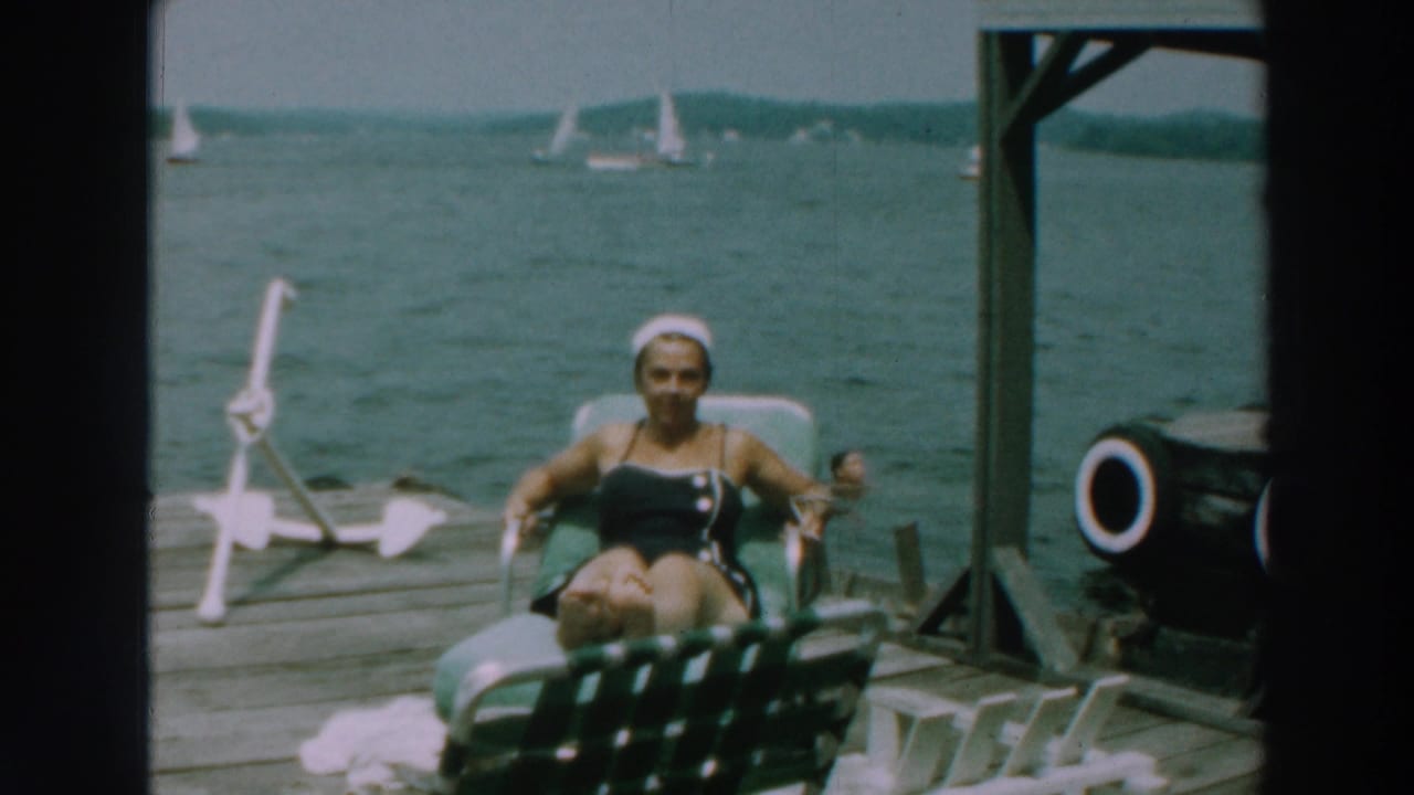 Woman in a dark swimsuit reclines on a green lounge chair on a wooden dock by the lake.