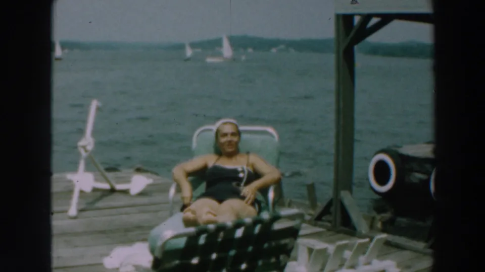 1964 Lake Winnebago home-movie frame of a woman smiling from a lounge chair on a wooden dock.