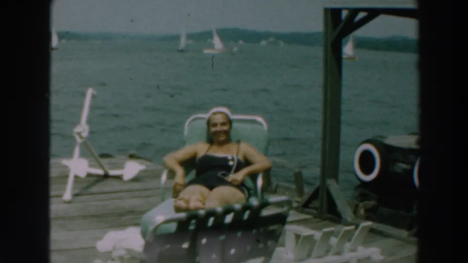 1964 Lake Winnebago frame showing dockside sunbathing, a lounge chair, lake water, and distant sailboats.