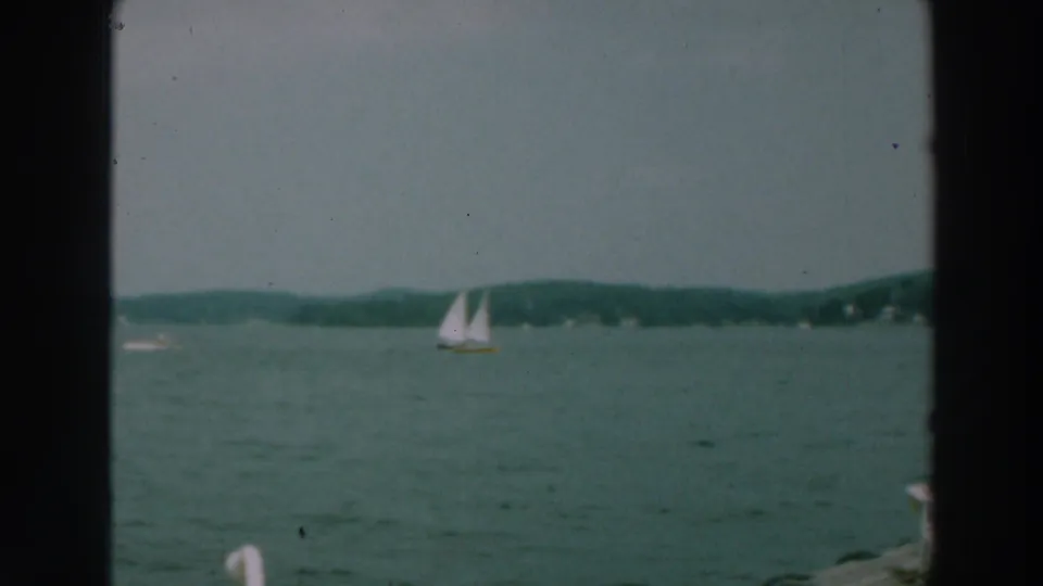 1964 Lake Winnebago 8mm frame with a distant white sailboat, low shoreline, green-blue water, and dark film borders.