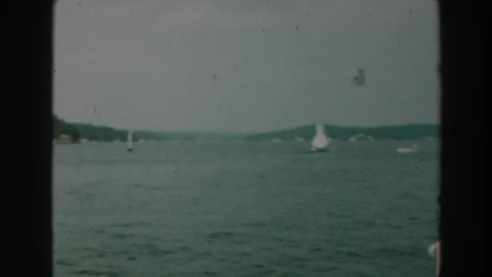 1964 home-movie frame from Lake Winnebago with small sailboats spread across the water and a soft wooded shoreline.