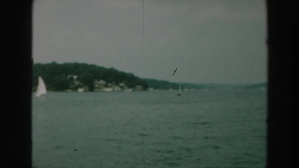 Lake Winnebago 8mm frame from 1964 with sailboats near a tree-covered shoreline and faint lakeside structures.