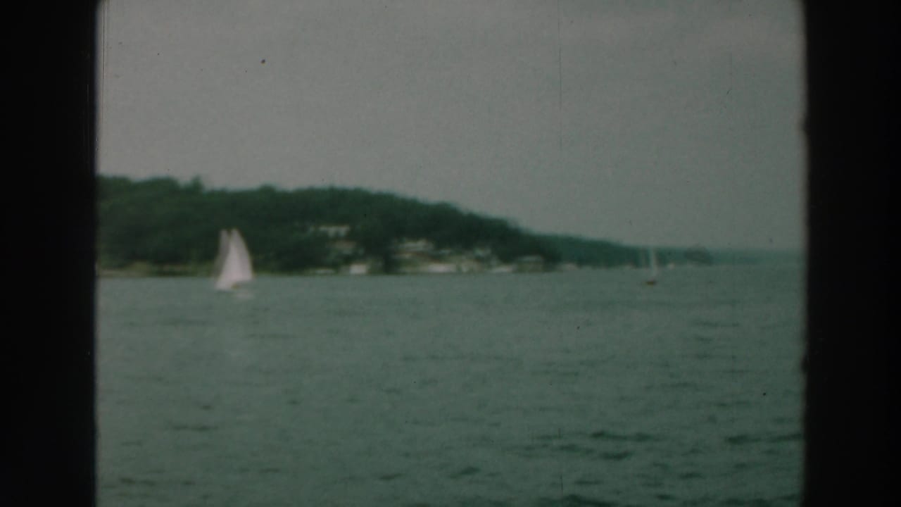 1964 Lake Winnebago view with a white sailboat crossing open water beneath a wooded Wisconsin shoreline.