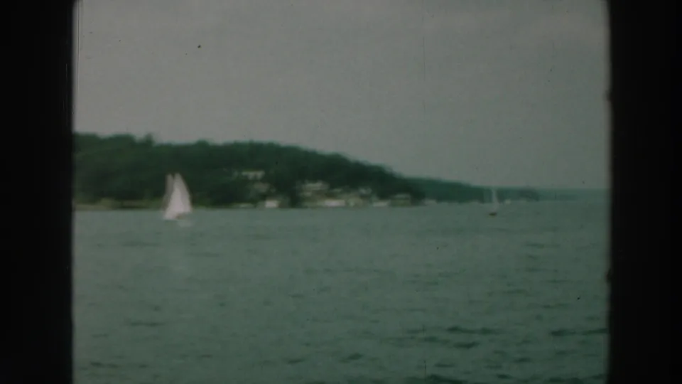 1964 Lake Winnebago view with a white sailboat crossing open water beneath a wooded Wisconsin shoreline.