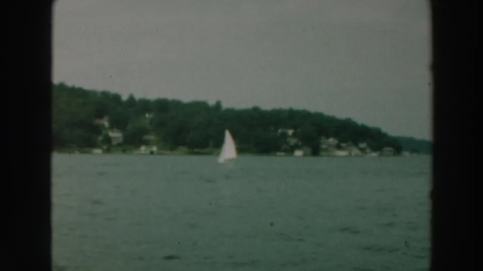 A white sailboat passes across Lake Winnebago in 1964, with distant houses set into the wooded shoreline.