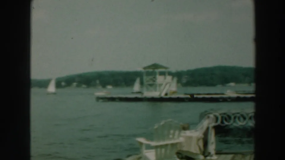 1964 Lake Winnebago dock view with white chairs in the foreground, a wooden pier, a tower, sailboats, and distant shoreline.
