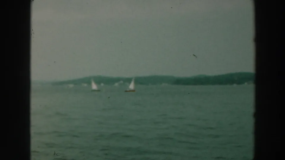 1964 Lake Winnebago home-movie frame showing two sailboats spread across calm water with wooded shore in the distance.