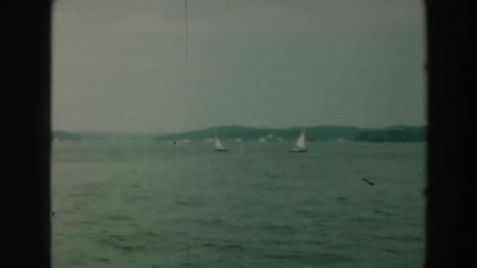 1964 Lake Winnebago frame with small distant sailboats, broad water surface, tree-lined horizon, and dark 8mm frame border.