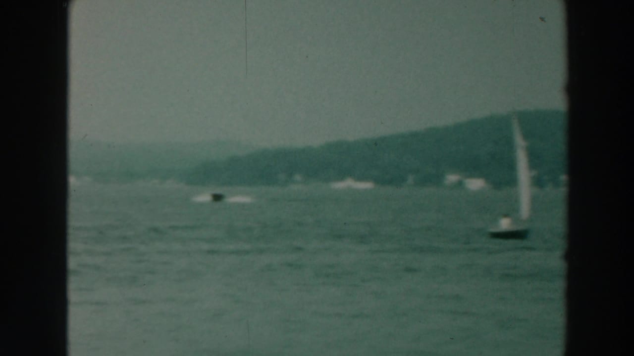 1964 8mm view of a small sailboat on Lake Winnebago with calm water, distant trees, and dark film-frame edges.