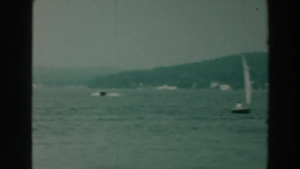 1964 8mm view of a small sailboat on Lake Winnebago with calm water, distant trees, and dark film-frame edges.