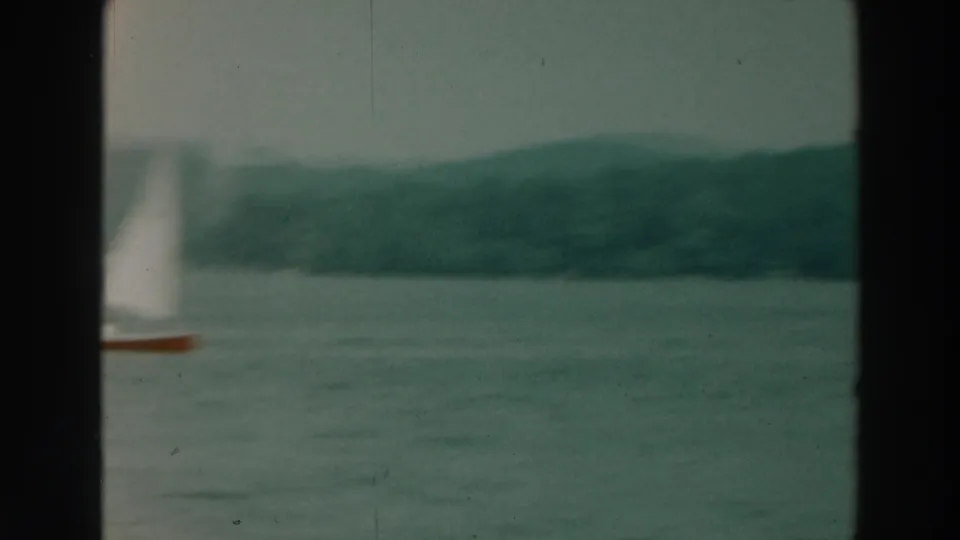 Soft 8mm frame of a white sailboat on Lake Winnebago, with muted water and a low forested shoreline behind it.