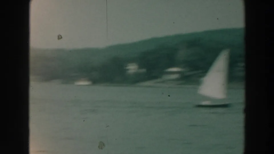 1960s home-movie frame showing a small sailboat on Lake Winnebago, filmed through soft focus with shoreline in the background.