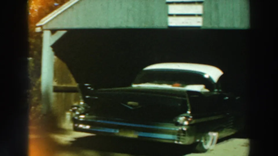 1964 Lake Winnebago frame with the rear of a dark car, white roof, tail fins, chrome bumper, and green garage siding.