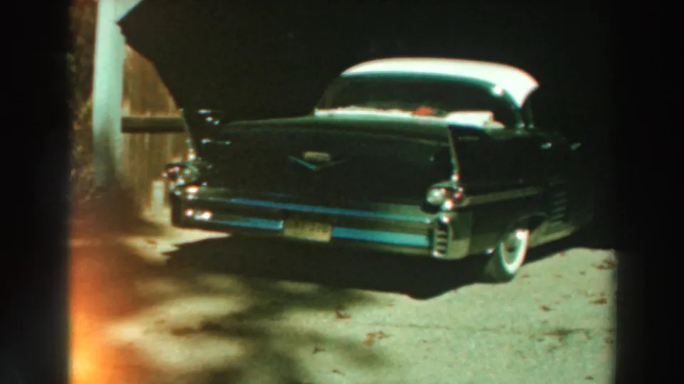 1964 Lake Winnebago home-movie frame showing a dark finned car from the rear side, with white roof, chrome details, and driveway light.