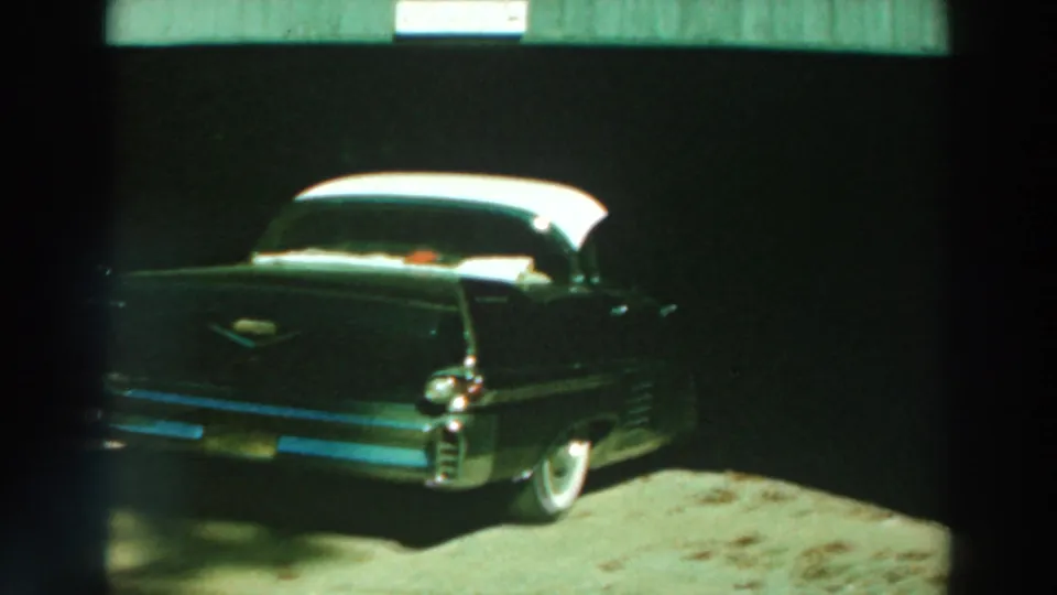 1964 Lake Winnebago frame showing a dark two-tone finned car in deep garage shade with chrome bumper and rear wheel visible.