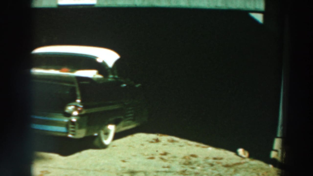 1964 Lake Winnebago home-movie frame showing a dark sedan with a white roof parked under a carport by a gravel drive.