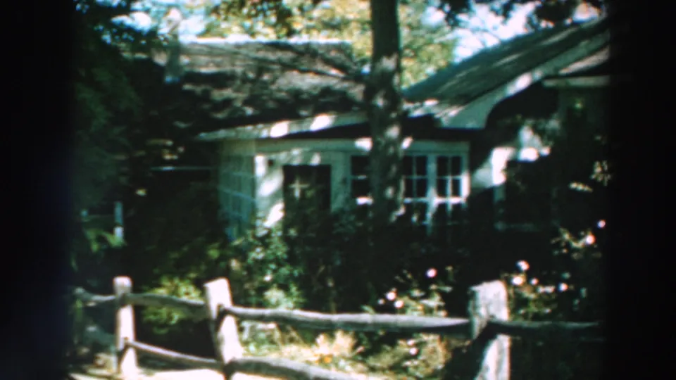Closer 8mm view of the yellow house, with white-trimmed windows behind shrubs and a split-rail fence in the foreground.
