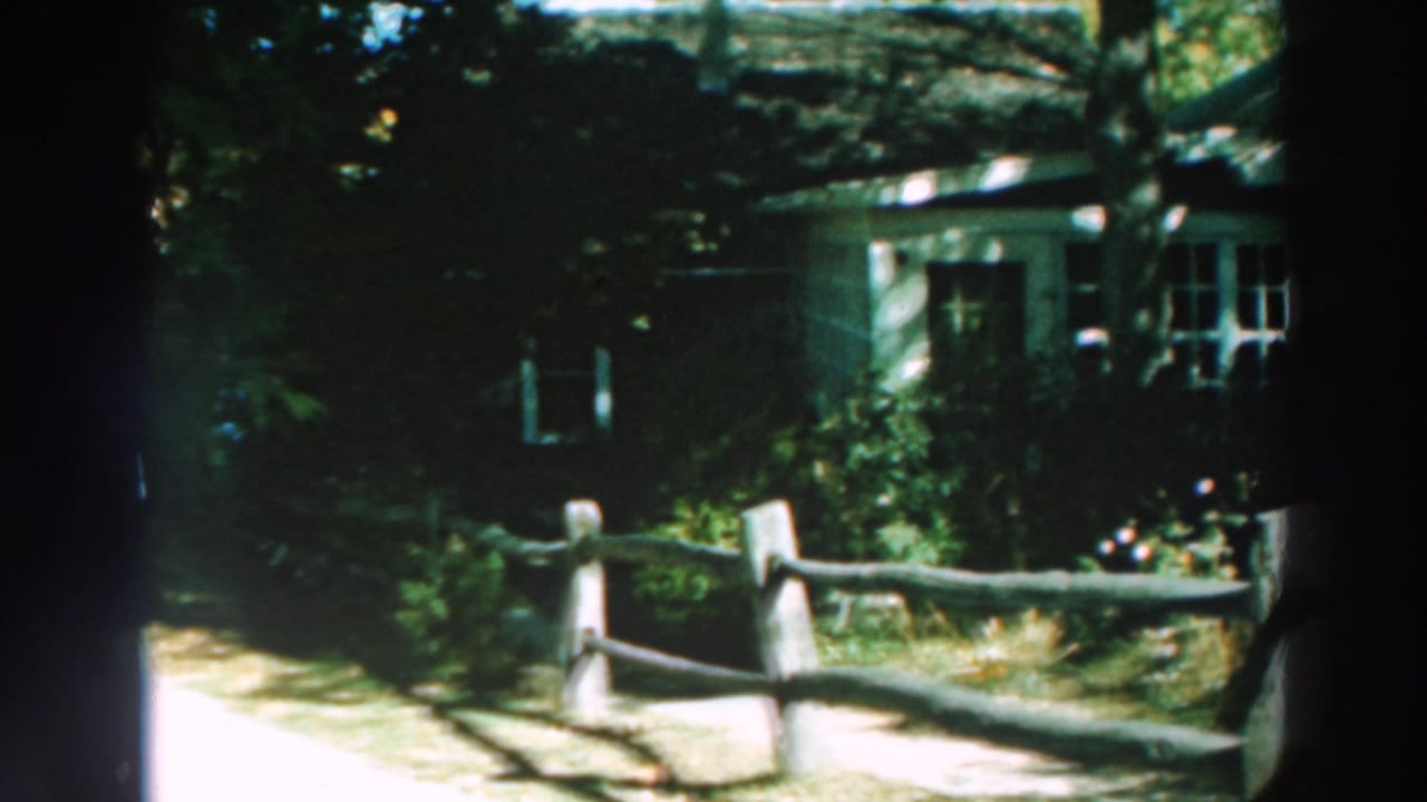 1964 Lake Winnebago home-movie frame showing a shaded house exterior, garden growth, split-rail fence, and bright path.