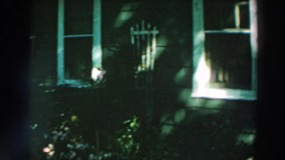 1964 Lake Winnebago home-movie frame showing dark siding, two windows, low garden plants, and deep shade across the facade.