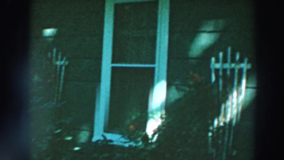 1964 Lake Winnebago home-movie frame with white house trim, garden stakes, shaded siding, and a bright patch of sunlight.