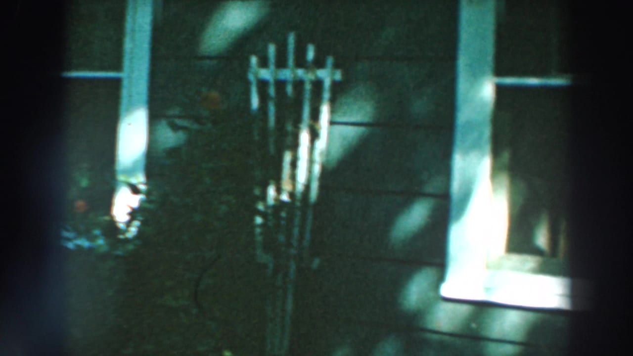 1964 Lake Winnebago home-movie frame showing wood siding, white windows, flowers, and shifting tree shadows.