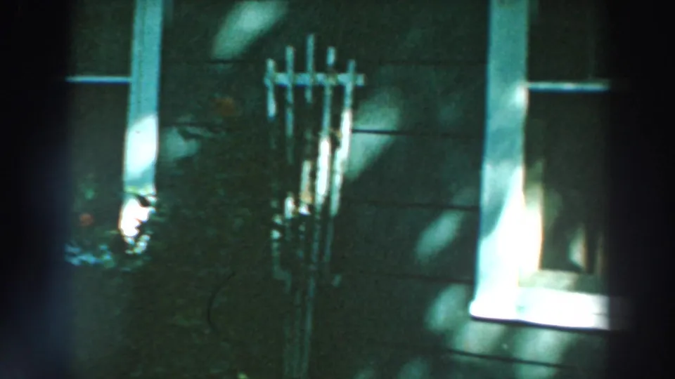 1964 Lake Winnebago home-movie frame showing wood siding, white windows, flowers, and shifting tree shadows.