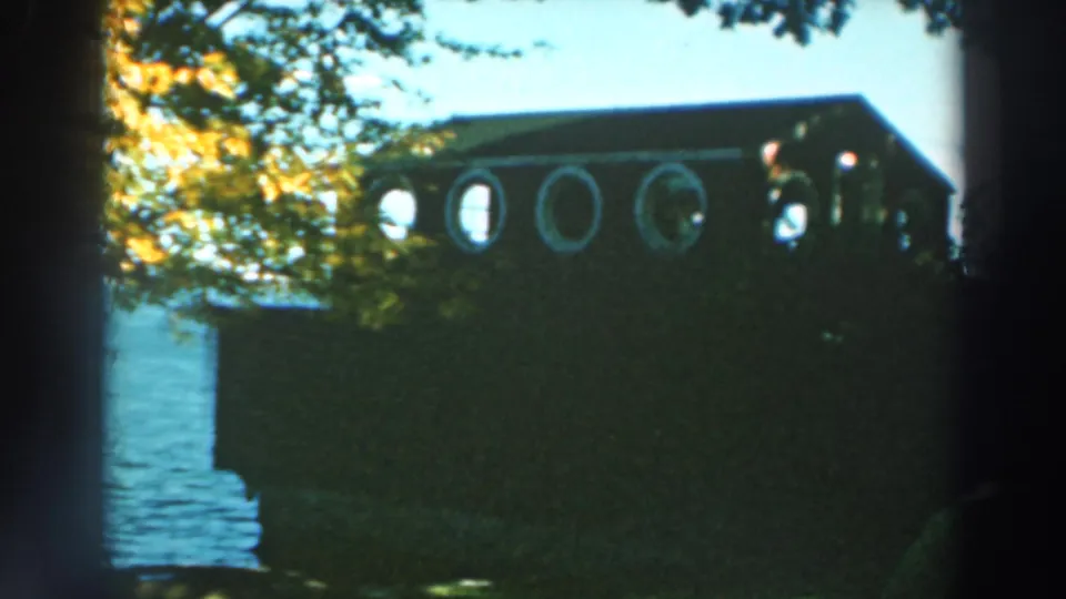 1964 Wisconsin home-movie frame of a wooden house with porthole-style windows near Lake Winnebago water and trees.