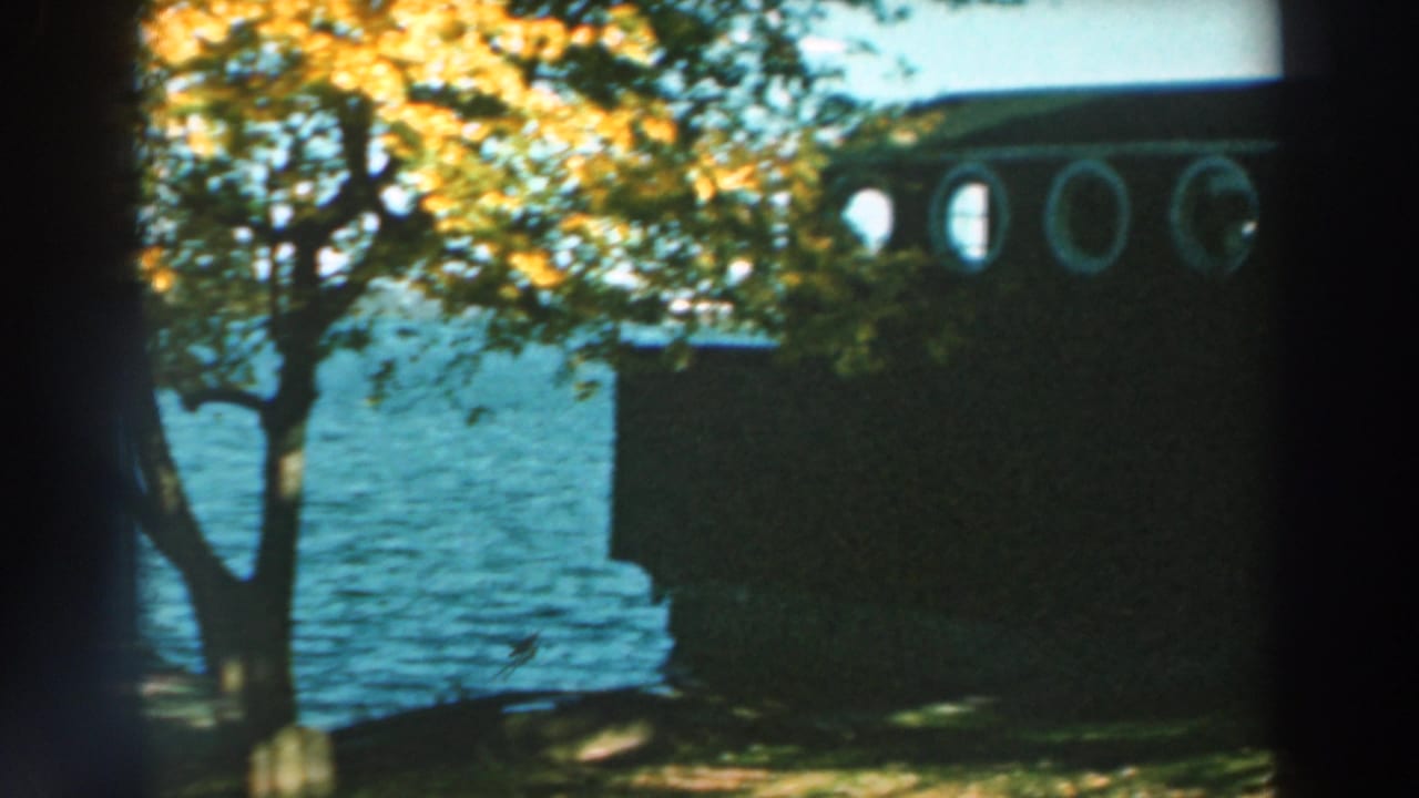1964 Lake Winnebago home-movie frame showing a dark waterfront building with circular windows, trees, and blue lake water.