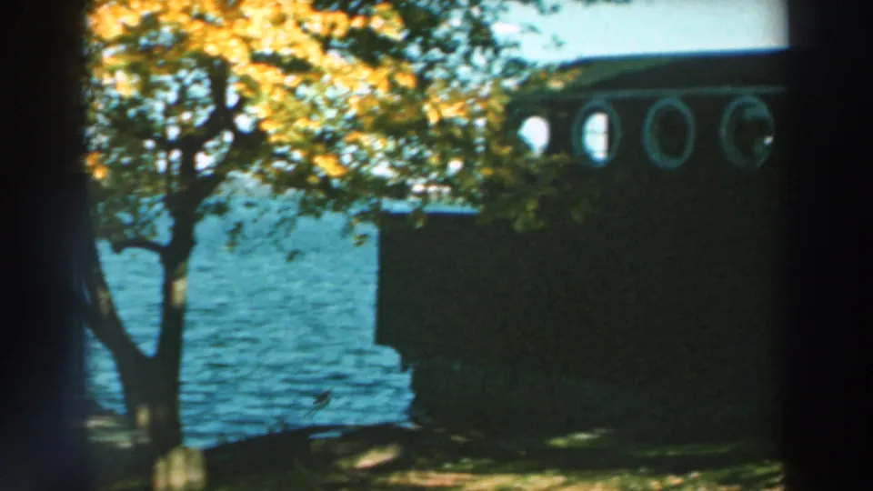 1964 Lake Winnebago home-movie frame showing a dark waterfront building with circular windows, trees, and blue lake water.