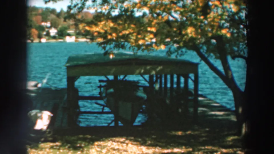 1964 Lake Winnebago frame with a covered dock, small boat, autumn tree canopy, blue water, and houses on the opposite shore.