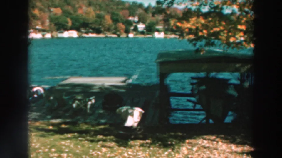 1964 Lake Winnebago shoreline view showing small boats near a covered dock, autumn leaves, open water, and distant houses.