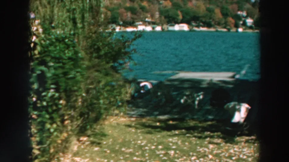 Lake Winnebago shoreline seen through trees, with blue water, a dock, boats, grass, and distant homes in a 1960s home-movie frame.